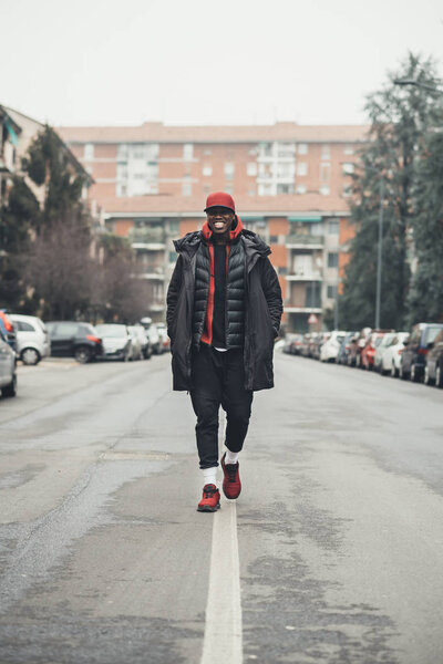 Молодой бородатый африканский мужчина на открытом воздухе ходит, глядя в камеру улыбается
