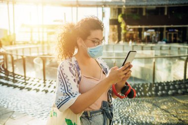 Açık havada akıllı telefon sohbeti yapan genç melez bir kadın. Cerrahi maske takan bir kadın. Virüsü ve kirliliği önlemek için cep telefonu kullanıyor.