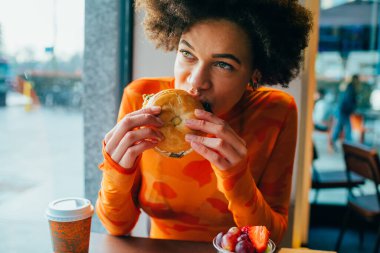 Genç ve aç bir afro kadın restoranda hamburger yiyor.
