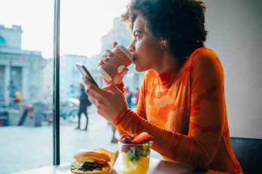 Genç afro kadın restoranda oturmuş akıllı telefon kullanarak kahve içiyor.