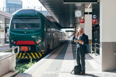 Demiryolu istasyonunda orta yaşlı bir işadamı tablet kullanarak akıllı telefonlardan konuşuyor - finans, ticaret, iletişim konsepti