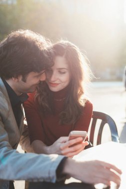 Dışarıda oturan kadın ve erkek akıllı telefon kullanıyor. Teknoloji trendlerini kullanan sevgi dolu bir çift. E-ticaret, oyun, konsept izleme.