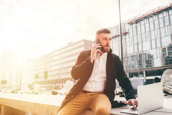 若いビジネスマンの屋外でコンピュータを使用してスマートフォンを話す ビジネスコールを持っている白人ひげそりの男リモートワーク ビジネス 作業概念 — ストック写真