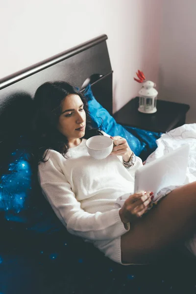 Young woman relaxing indoors reading using tablet - Female at home watching movie having cup of hot drink - coziness, entertainment, technology concept