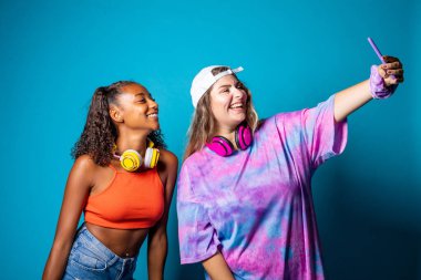 Mavi arka planda giyinmiş iki genç arkadaş selfie çekiyorlar - akıllı telefon kullanarak izole edilmiş farklı kadın canlı yayın - vlog, canlı yayın, sosyal ağlar konsepti