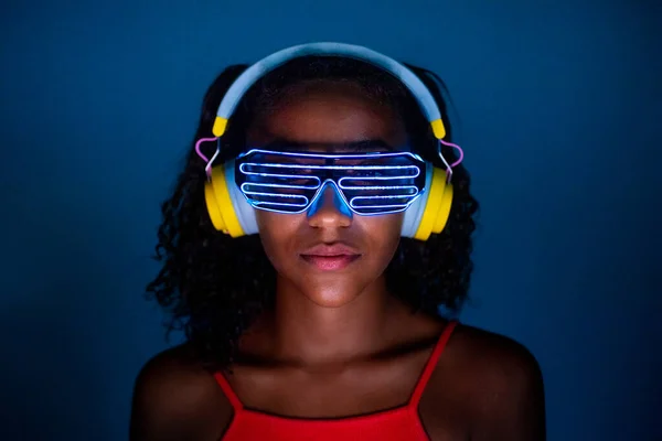 Young woman wearing headphones and futuristic led glasses on blue background - Isolated black woman wearing 3d smart glasses and headphones - virtual reality, future, technology concept