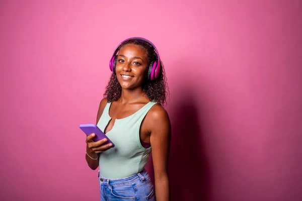 ピンクの背景で陽気な黒人女性が音楽を聞いているスマートフォンを保持 ヘッドフォンを身に着けている電話を使用して独立した黒の若い女の子 ストリーミング 音楽コンセプト — ストック写真