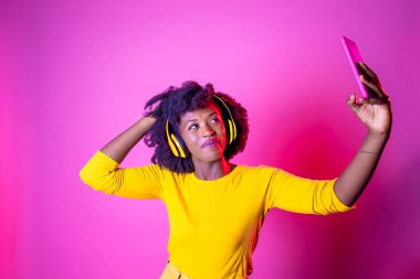 Pembe arka planda selfie çeken genç ve güzel bir zenci kadın - izole edilmiş Afrikalı genç bir kadın canlı yayında kulaklık takıyor stüdyo fotoğrafı - sosyal ağ, teknoloji, çeşitlilik konsepti