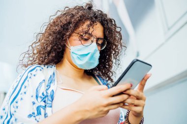Çok ırklı, koruyucu maske takan, cep telefonu kullanan, açık havada poz veren genç bir kadın, şehirde akıllı telefon kullanan karışık ırk genç kadın, koruma, kirlilik, teknoloji kavramı.