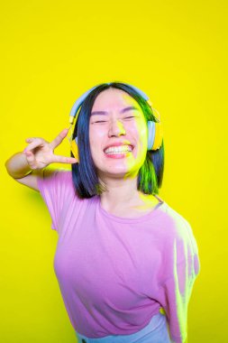 Gülümseyen genç kadın sarı arka planda izole edilmiş müzik dinliyor kulaklıkla müzik dinleyen Asyalı genç kız zafer işareti gösteriyor başarı, pozitif, zafer konsepti