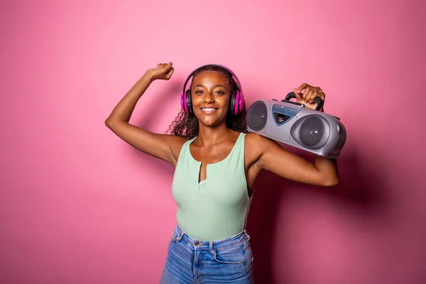 Young beautiful black woman dancing indoor on pink baground holding vintage stereo - Isolated diverse female clubbing holding vintage radio - happiness, excitement, dancing concept