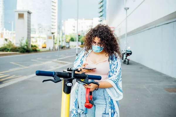 Young multiethnic woman wearing mask unlocking escooter - Diverse ecological female outdoor using smartphone driving kickscooter - health care, ecological, technology concept