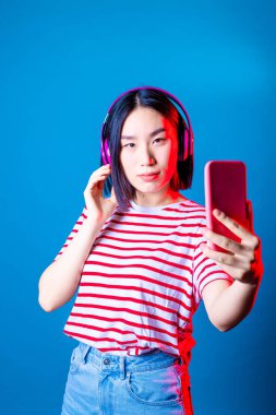 Genç ve güzel Asyalı bir kadın mavi arka planda selfie çekiyor - izole edilmiş Asyalı genç bir kadın canlı yayında kulaklık takıyor stüdyo fotoğrafı - sosyal ağ, teknoloji, çeşitlilik konsepti