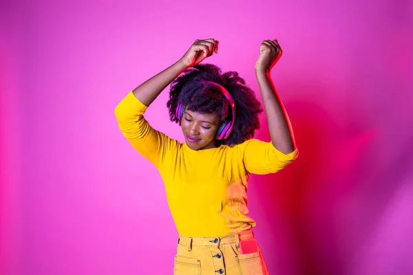 Young beautiful black woman dancing indoor on pink baground holding smartphone - Isolated diverse female clubbing listening streaming music - happiness, excitement, dancing concept