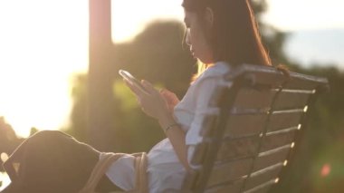 Genç bir kadın parktaki bankta oturur ve güzel bir sabah güneşinde akıllı telefon kullanır..