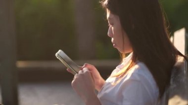 Genç bir kadın parktaki bankta oturur ve güzel bir sabah güneşinde akıllı telefon kullanır..