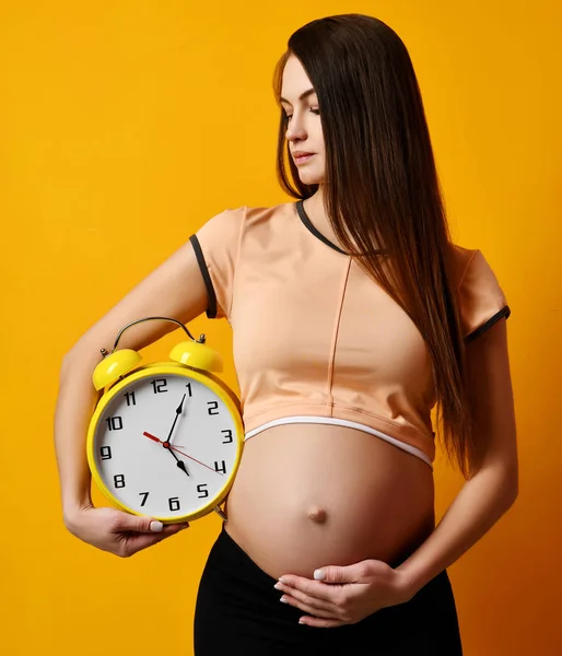 Zwangere vrouw in top shirt houden grote klok horloge glimlachend op geel — Stockfoto