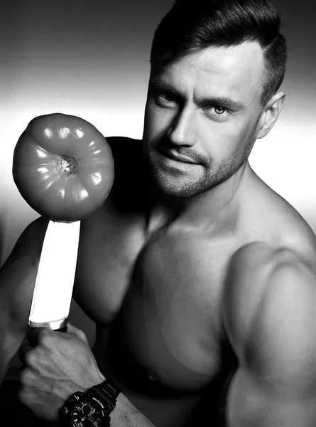 Muscular sport man standing on kitchen and hold big knife with tomato — Stock Photo, Image