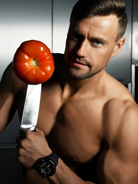 Muscular sport man standing on kitchen and hold big knife with tomato — Stock Photo, Image