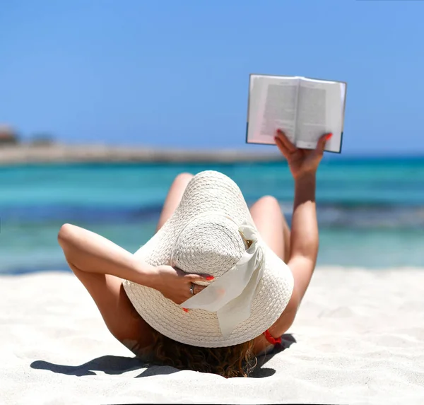 Girl lying on sand sea shore beach in white summer hat reading book — Stock Photo, Image