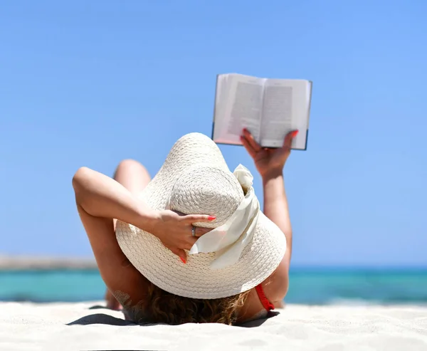 Girl lying on sand sea shore beach in white summer hat reading book — Stock Photo, Image