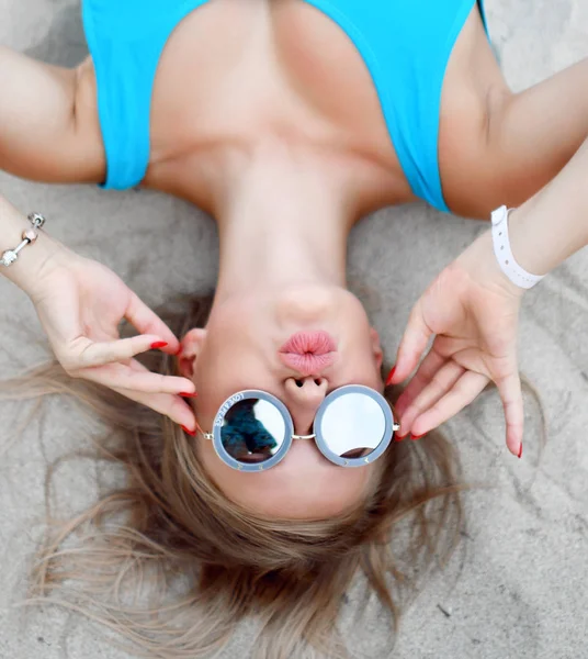 Young beautiful blonde girl lying on sand tropical on tropical beach in blue body vest and round sunglasses kissing — Stock Photo, Image
