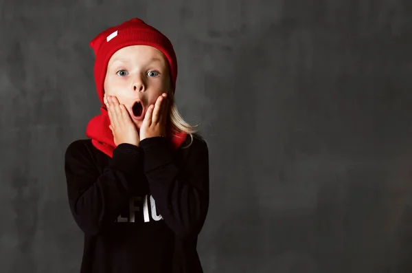 Niña pequeña niña posando en sombrero rojo y bufanda sorprendida gritando en gris oscuro — Foto de Stock