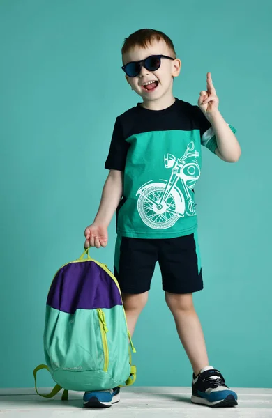 Niño preescolar de pie con mochila y gafas de sol en pantalones cortos y camiseta en menta verde —  Fotos de Stock