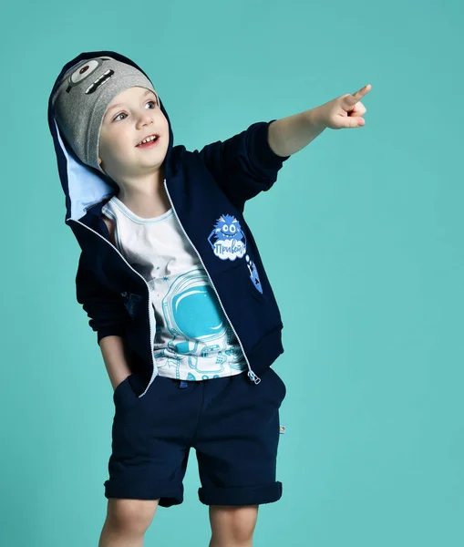 Chłopiec dziecko stojąc w niebieska Bluza zip-up i szary kapelusz w krótkich spodenkach, wskazując ręką na zielonej mięty — Zdjęcie stockowe