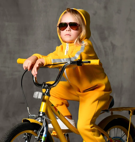 Jong meisje kind rit zitten op geel fiets in zonnebril op grijs betonnen muur opzoeken — Stockfoto