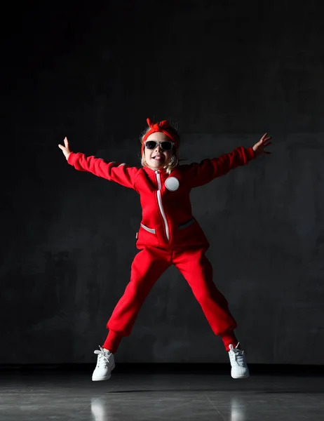 Niña pequeña saltando en tela roja y gafas de sol en la pared gris oscuro — Foto de Stock