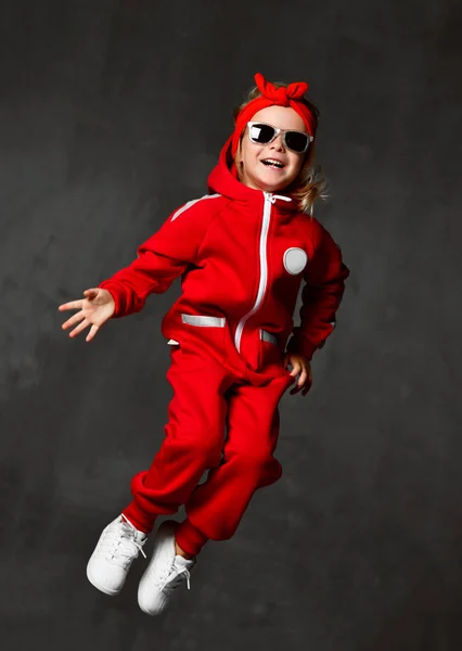 Genç kız çocuk kırmızı kumaş ve güneş gözlüğü koyu gri duvara atlama bebek — Stok fotoğraf