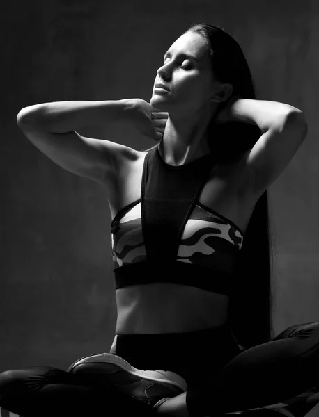 Молода спортивна жінка в тренувальній тканині позує, розтягуючись в тренажерному залі на темно-сірому фоні — стокове фото