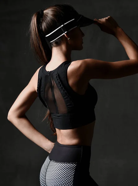 Mujer deportiva joven en paño de ejercicio y gorra posando estirándose en el gimnasio sobre fondo gris oscuro — Foto de Stock