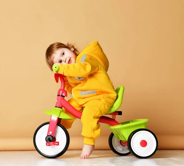 Baby girl kid toddler riding her first bicycle tricycle in warm yellow overalls looking up on warm brown — Stock Photo, Image