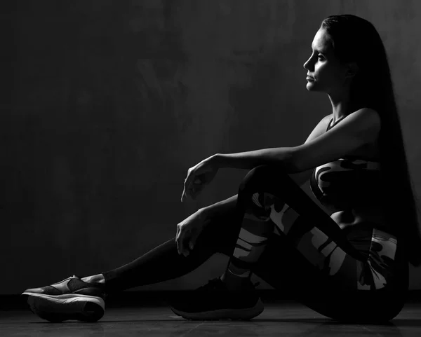 Junge Sportlerin in Trainingsjacke posiert sitzend Stretching im Fitnessstudio auf dunkelgrau — Stockfoto