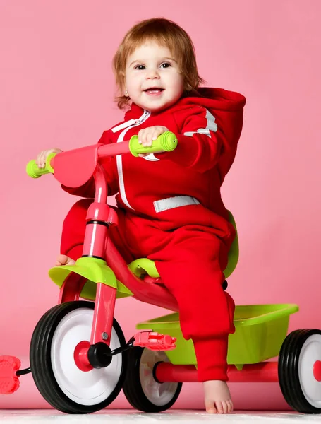 Baby girl kid riding her first bicycle tricycle in red overalls looking up on light pink — Stock Photo, Image