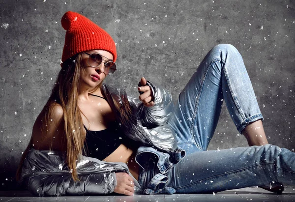 Young sport woman after workout exercise sitting point finger in silver sexy thin down puffer jacket blue jeans red hat