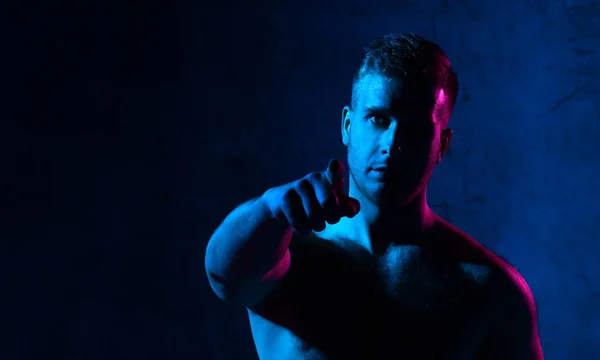 Неоновый светлый портрет сексуального улыбающегося мужчины с мускулистым телом, указывающим пальцем на темноту — стоковое фото