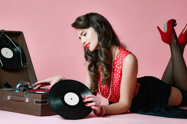 Гламурная девушка-пинап лежит рядом с ретро-граммофоном, держа пластинку с пластинкой, глядя на розовый фон. — стоковое фото