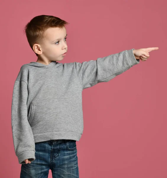 Niño en sudadera gris apuntando con un dedo a la esquina sonriendo aislado en rosa —  Fotos de Stock