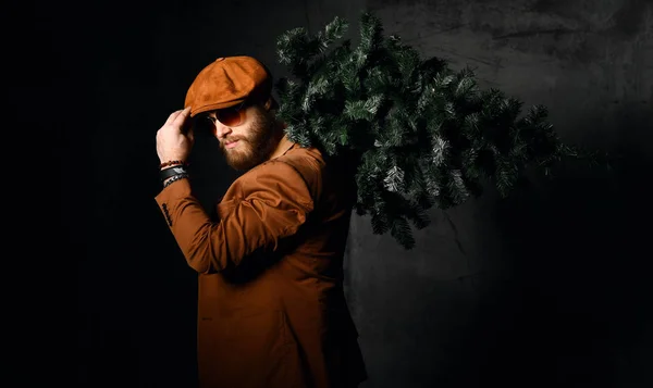 Жорстокий бородатий чоловік в коричневій шкіряній куртці і капелюсі авіатор сонцезахисні окуляри тримають різдвяну ялинку — стокове фото