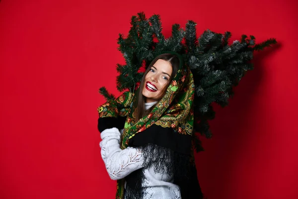 Happy Russian style woman hold Christmas  tree on her shoulders happy smiling laughing on dark red background