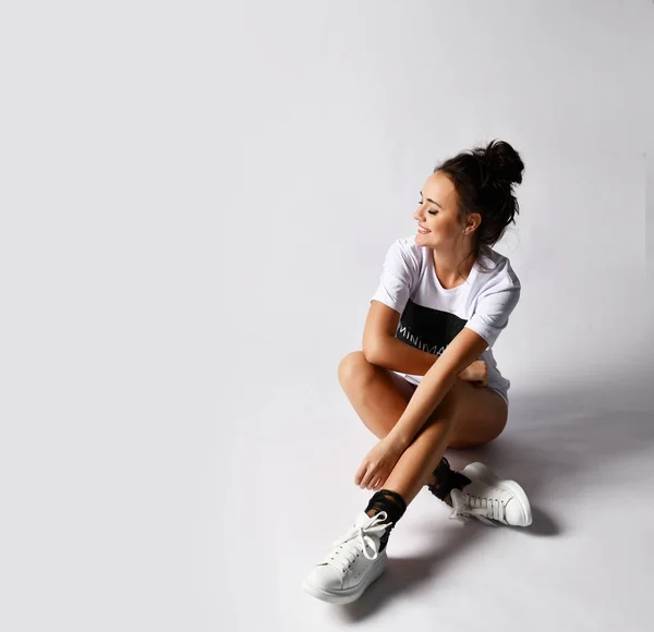 Hermosa mujer morena sentada sobre fondo gris en larga camiseta blanca y zapatillas de deporte —  Fotos de Stock