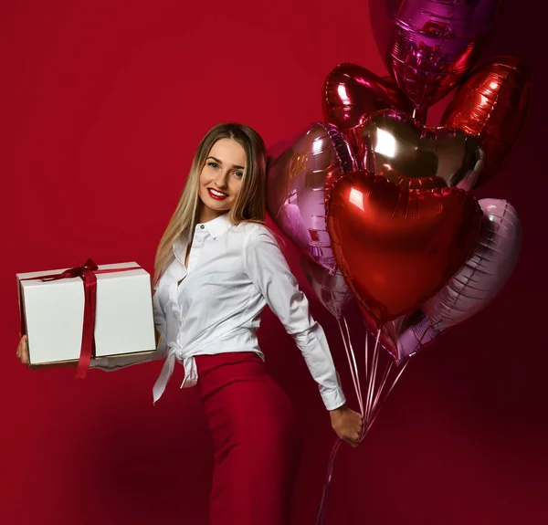 Mooie vrouw met huidige geschenk doos en paars roze hart ballonnen gelukkig lachend op rode achtergrond — Stockfoto