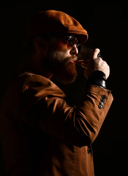 Brutal hombre de negocios con barba y bigote en gorra marrón y chaqueta beber whisky —  Fotos de Stock