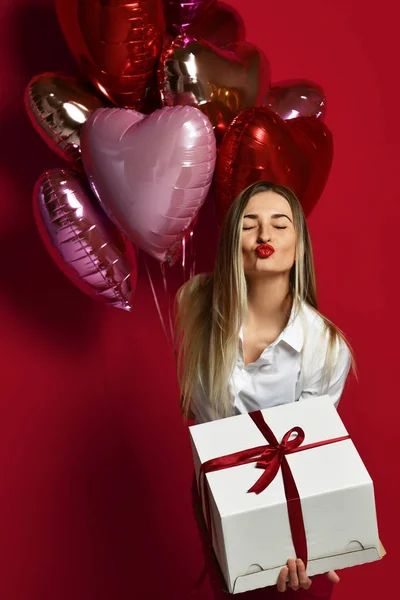 Mooie vrouw met huidige geschenk doos en paars roze hart ballonnen gelukkig waait kus op rode achtergrond — Stockfoto