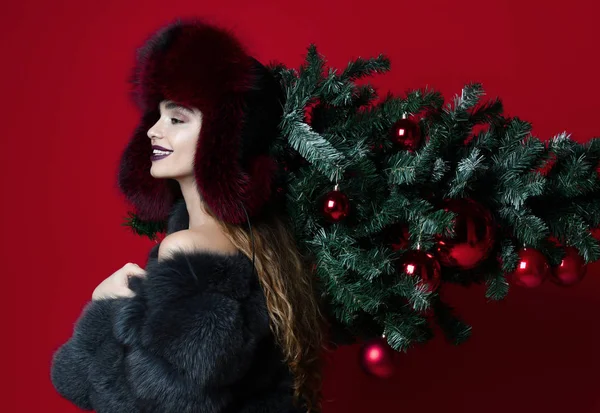 Glada mode kvinna i vinter mössa och päls kappa håller inredda xmas tree på axlarna leende — Stockfoto