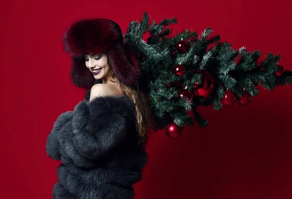 Feliz mujer de la moda en el sombrero de invierno y abrigo de piel sostienen árbol de Navidad decorado en sus hombros sonriendo —  Fotos de Stock