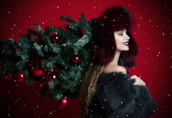 Feliz mujer de la moda en el sombrero de invierno y abrigo de piel sostienen árbol de Navidad decorado en sus hombros sonriendo — Foto de Stock